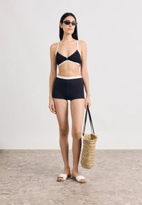 Μαύρο μπικίνι με μπεζ λεπτομέρειες, σορτς ψηλόμεσο και κοντό τοπ. Συνδυασμένο με γυαλιά ηλίου και πλεκτή τσάντα ώμου.