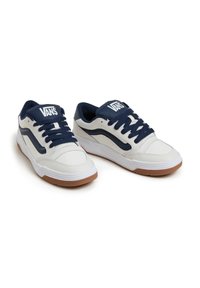 Valkoiset nahkakengät, joissa on tumman siniset yksityiskohdat ja nauhat, ja joissa on teksturoitu kengän malli sekä kumipohja. Vans-logo kielen kohdalla.