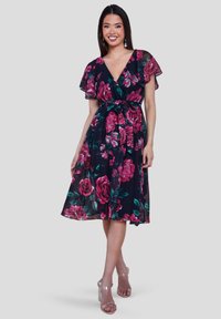 Wanita mengenakan gaun hitam panjang lutut dengan motif bunga pink, lengan melayang, leher V, dan ikat pinggang, dipadukan dengan sandal hak tinggi bening.