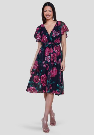 Wanita mengenakan gaun hitam panjang lutut dengan motif bunga pink, lengan melayang, leher V, dan ikat pinggang, dipadukan dengan sandal hak tinggi bening.