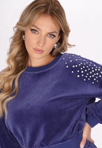 Lila sammetsurtröja med rund halsringning, dekorerad med spridda rhinestones på ena axeln, med ribbade ärmslut och midja.