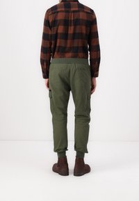 Zelené cargo kalhoty s bočními kapsami, elastickým pasem a zúženými nohavicemi, spojené s hnědým a černým kostkovaným košilí. Tkanina vypadá jemně.