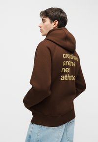 Junger Mann mit kurzen dunklen Haaren, trägt einen braunen Hoodie mit dem Text "creatives are the new athlete" auf dem Rücken und eine hellblaue Jeans.