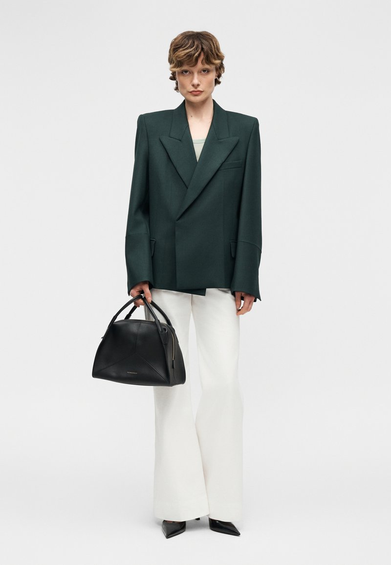 Personne portant un blazer oversize vert foncé, pantalons larges blancs, chaussures pointues noires, tenant un sac à main structuré noir sur un fond blanc.