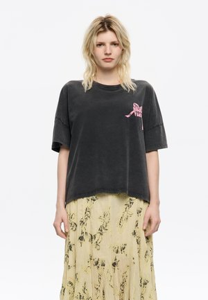 T-shirt nera oversize in cotone con maniche corte e un design grafico rosa sul lato sinistro del petto, indossata sopra una gonna gialla a fantasia floreale.