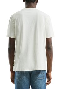 Man in een eenvoudig wit T-shirt met korte mouwen en blauwe denim jeans, gezien vanuit de achterkant tegen een witte achtergrond.