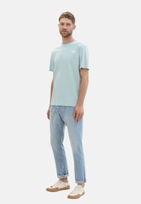 Helles blaues Baumwoll-T-Shirt mit kurzen Ärmeln und Rundhalsausschnitt, kombiniert mit hellen Jeans und beigen Sneakers in niedrigem Schnitt mit braunen Akzenten.