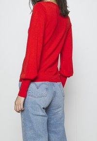 Roter gestrickter Pullover mit langen, puffigen Ärmeln und einem taillierten Saum, kombiniert mit hellblauen hochgeschnittenen Jeans.