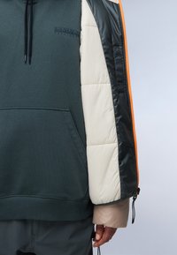 Hoodie met een donkergroene romp, romige puffy mouwen, glanzende zwarte accenten, een oranje rits en een voorste kangoeroezak. Voorzien van verstelbare manchetten.