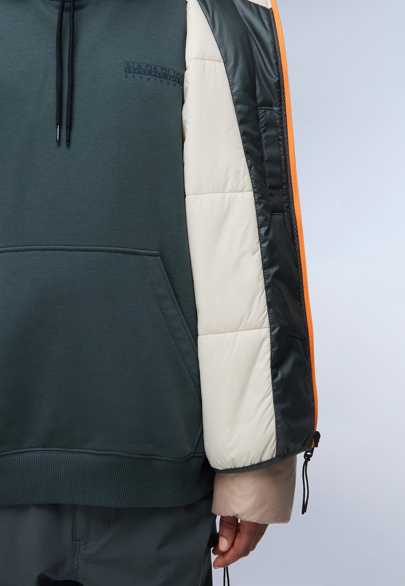 Sudadera con un cuerpo verde oscuro, mangas abullonadas color crema, detalles en negro brillante, cremallera naranja y un bolsillo tipo canguro en la parte frontal. Cuenta con puños ajustables.