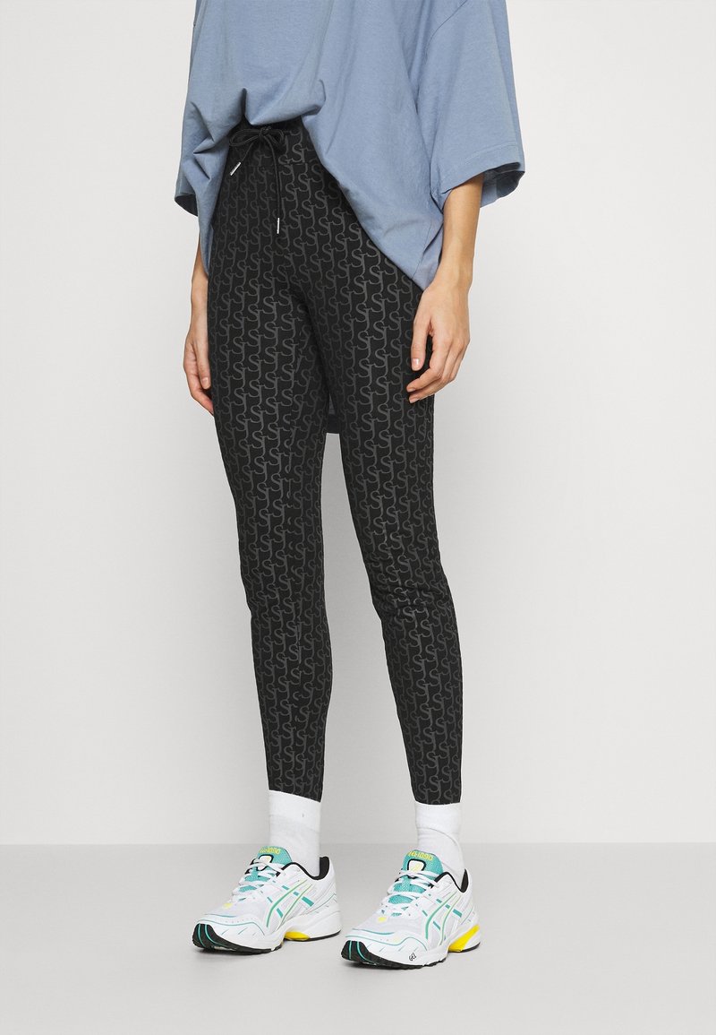 Svarta leggings med ett grått mönster, med dragsko i midjan. Kombineras med vita sneakers med turkosa och gula detaljer.