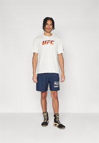 Bela kratka majica s kratkimi rokavi z rdečim logotipom "UFC", v kombinaciji z mornarsko modrimi kratkimi hlačami z grafičnim potiskom in črnimi borbenimi škornji.