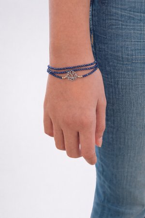 Hand trägt ein blaues Wickelarmband mit Perlen und silbernem Lotus-Anhänger, ruht neben einem Bein in blauen Jeans vor weißem Hintergrund.