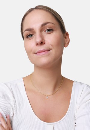 Goldene Halskette mit feiner Kette und kleinem Tropfenanhänger, getragen von einer Person mit einem hellen, gerippten Oberteil. Hautfarbe ist natürlich.