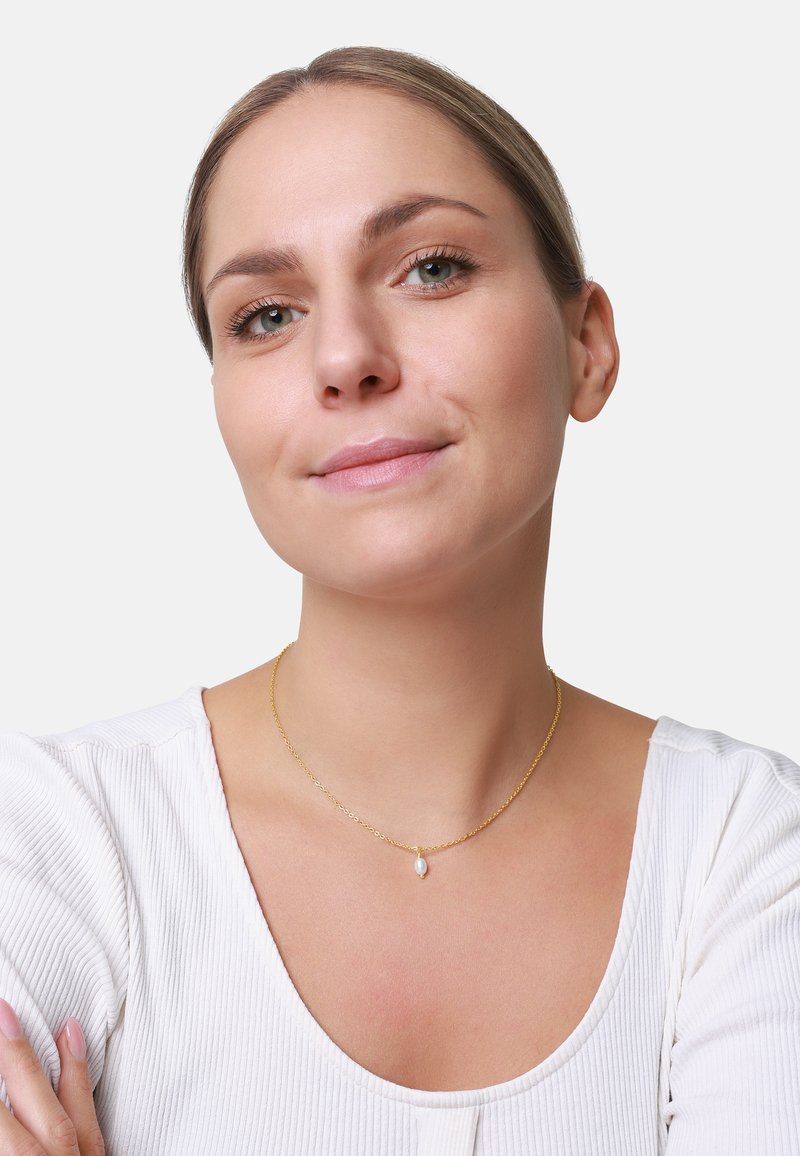 Collier en or avec une chaîne délicate et un petit pendentif en forme de goutte, porté par une personne avec un haut rayé clair. Le teint est naturel.