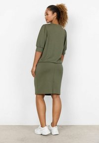 Vestido verde oliva con mangas cortas abullonadas, textura acanalada y silueta ajustada, combinado con zapatillas blancas. Se muestra la vista trasera.