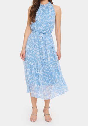 Mujer con vestido midi sin mangas de patrón azul y blanco, con cinturón en la cintura y escote con volantes, combinado con tacones con tiras.