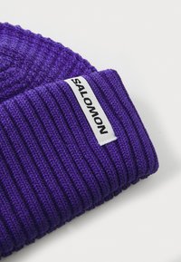 Paarse gebreide muts met ribbeltextuur. Heeft een wit label met zwarte tekst "SALOMON" op de omgevouwen rand.