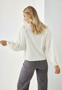 Blusa blanca de lino con mangas largas y abullonadas, decorada con un borde de encaje. Combinada con pantalones grises de corte relajado. Diseño simple y limpio con una textura ligera.