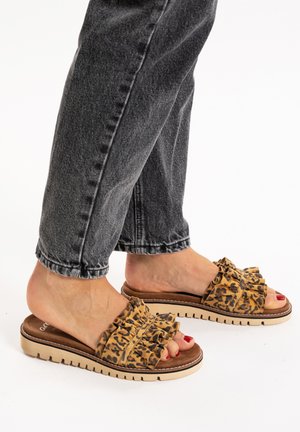 Infradito con stampa leopardata e dettagli a volant, di colore beige, con suola imbottita. Indossati con jeans grigi scuri risvoltati. Smalto rosso sulle unghie dei piedi visibile.