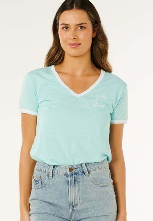 Femme aux longs cheveux bruns portant un t-shirt bleu clair à col en V avec un bord blanc et un jean taille haute bleu clair, debout devant un fond uni.