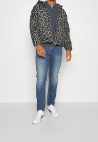 Chaqueta con cremallera de estampado de leopardo en gris y negro, combinada con un polo oscuro, vaqueros azules y zapatillas blancas con detalles en azul marino.