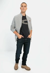 Veste bomber matelassée grise sur un T-shirt noir avec un grand logo, accompagnée de jeans foncés et de bottes lacées couleur fauve. Design et coupe décontractés.