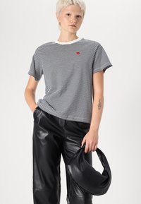 T-shirt rayé noir et blanc avec un logo en forme de cœur rouge, associé à un pantalon en cuir noir taille haute. Le mannequin tient un sac noir courbé.