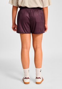 Kort, mörklila shorts med elastisk midja och slät textur. Bakifrån syns en skräddarsydd passform och minimalistisk design. Bärs med vita strumpor och skor.