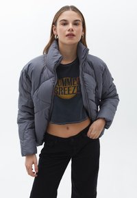 Chaqueta acolchada gris con un cuello alto, que presenta un diseño acolchado. Combinada con una camiseta gráfica corta y pantalones negros de tiro alto.