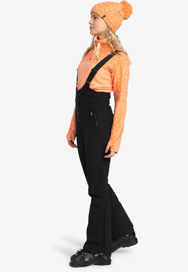 Oranje gestreepte blouse met lange mouwen, zwarte tuinbroek met verstelbare banden en zwarte snowboots. Bevat ritszakken aan de zijkant en een getextureerde gebreide muts.
