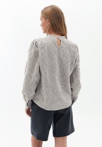 Camicia grigia con texture e motivo a foglia, maniche a sbuffo e dettaglio a chiave sul retro, abbinata a pantaloni corti neri. Leggera lucentezza e tessuto morbido.