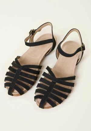 Sandales noires à lanières avec une tige douce et texturée, un design à bout ouvert, une bride réglable à la cheville, et une semelle plate couleur tan.