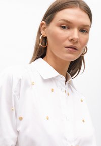 Camicia bianca dal taglio rilassato, con decorazioni dorate e colletto classico. Il modello indossa grandi orecchini a cerchio dorati.