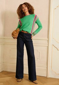 Zaļš adīts džemperis ar rozā un purpura svītrām, apvienots ar tumšiem platām kājām džinsiem. Modelis tur brūnu somu, stāvot uz koka grīdas.