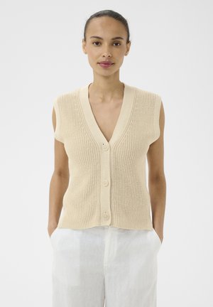 Kvinde iført ærmeløs beige strikket cardigan med knapper og hvide bukser, stående med hænderne i lommerne mod en ensfarvet baggrund.