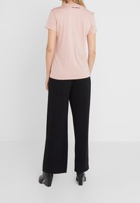 Vaaleanpunainen puuvillainen t-paita, jossa on lyhyet hihat ja pyöreä kaula-aukko. Yläselässä on musta logo. Yhdistetty mustiin, leveälahkeisiin housuihin.