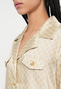 Béžová hodvábna košeľa s bielym monogramovým vzorom, vybavená vrchným golierom, zlatými gombíkmi a nádobkami na prsia.