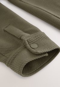 Tessuto verde oliva con texture a righe, dotato di un polsino arrotolato e un dettaglio di bottone scuro sulla manica.