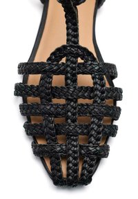Sandal de cuero trenzado negro con punta abierta, que presenta un patrón de trenzado entrecruzado y una suela beige. Diseño plano sin tacón.