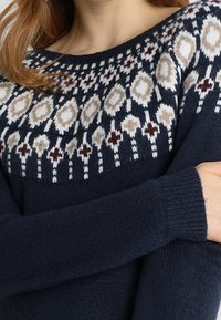 Femme portant un pull bleu marine avec un motif géométrique beige, blanc et maroon sur le haut de la poitrine et les épaules.