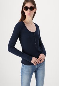 Mornarska majica dugih rukava typu henley, rebrasta tkanina, sadrži šest dugmadi sprijeda, u kombinaciji s svijetloplavim trapericama i crnim naočalama u obliku mačjih očiju.