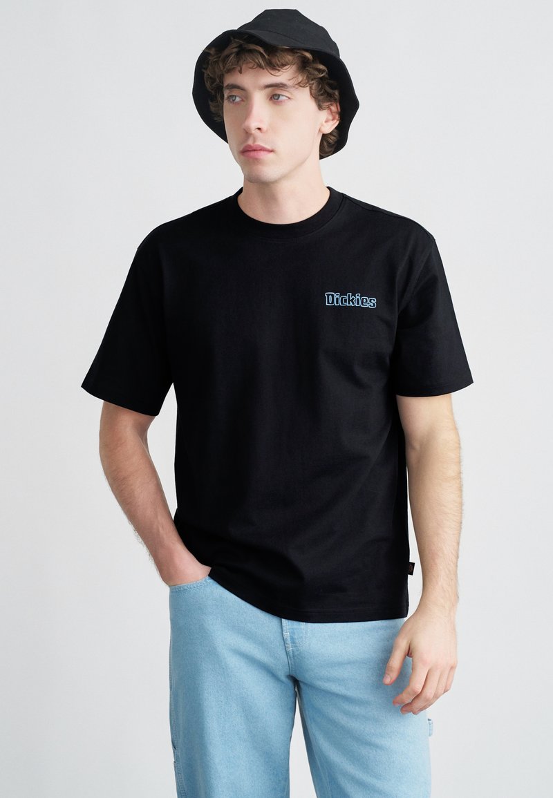 Μαύρο βαμβακερό μπλουζάκι με κοντά μανίκια, με μπλε λογότυπο «Dickies» στο αριστερό μέρος του στήθους. Συνδυάζεται με τζιν παντελόνι ανοιχτού μπλε χρώματος.