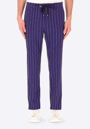 Pantaloni blu navy con sottili righe verticali beige, coulisse nera regolabile, chiusura con bottoni, indossati con sneakers bianche e camicia infilata dentro.