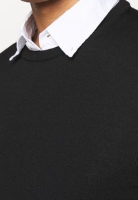 Pull en tricot noir avec un col ras du cou, superposé à une chemise blanche à col. La texture est lisse et ajustée, mettant en valeur la zone du cou.