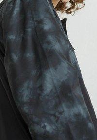 Abrigo negro con un patrón de tie-dye en gris oscuro en las mangas. Textura suave con un ligero brillo. Detalles de diseño minimalistas.
