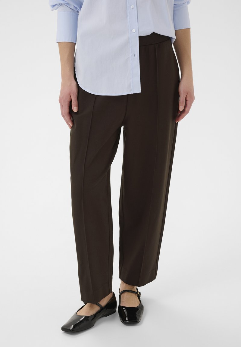 Personne portant un pantalon marron foncé ample, une chemise bleu clair à manches longues boutonnée et des ballerines Mary Jane noires, debout sur un fond blanc.
