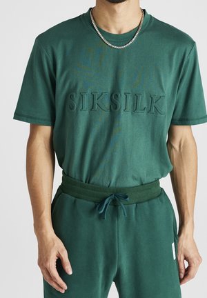 Ciemnozielony t-shirt z krótkim rękawem z wytłoczonym logo „SIKSILK”, zestawiony z dopasowanymi joggerami mającymi ściągacz z regulowanym sznurkiem w pasie oraz kieszenie.