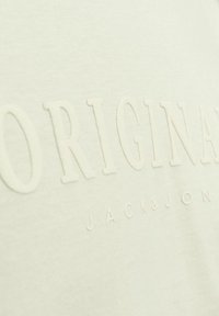Cămașă tip T de culoare bej deschis, confecționată din bumbac, cu inscripții în relief "ORIGINAL" și "JACK&JON". Textură netedă cu un design casual.