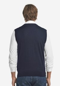 Marinblå ärmlös tröja i mjukt material, med rundad halsringning och ribbad nederkant, i kombination med en vit långärmad skjorta under.